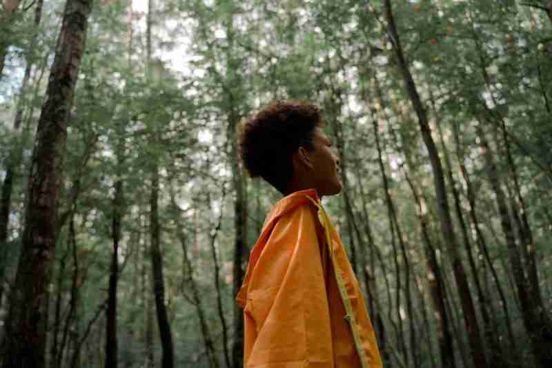 Lady in orange wrap and blown out afro hair standing among tall green trees in a state of contemplation  of her feelings of anxiety