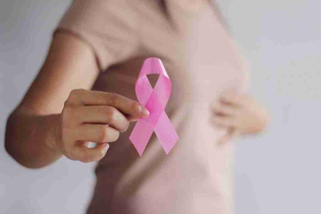 Lady in a beige top holding a pink ribbon in one hand and touching her chest with the other for breast cancer risks