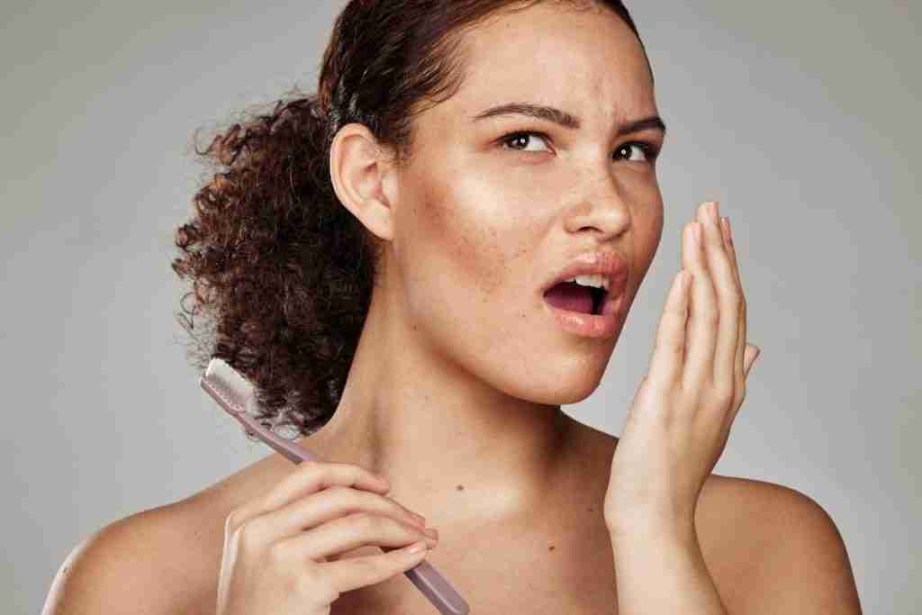 Pretty lady holding a toothbrush but sniffing her breath - bad breath is one of the Major Dental Warning Signs