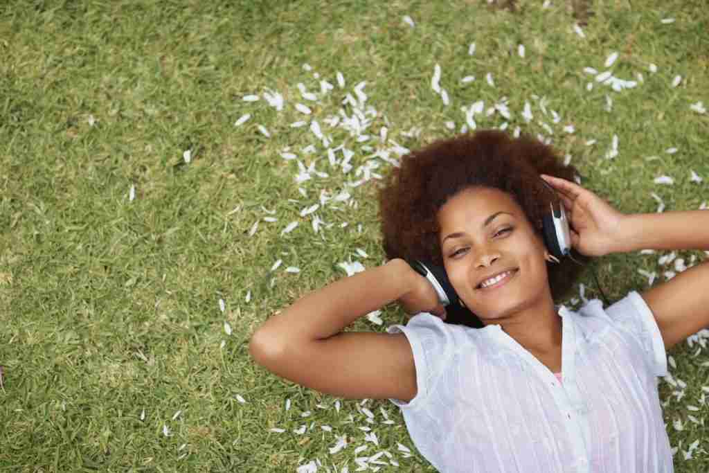 Black woman lying on green grass outdoors smiling with headphones on - Nature and Music are Therapies for Powerful Self-Care