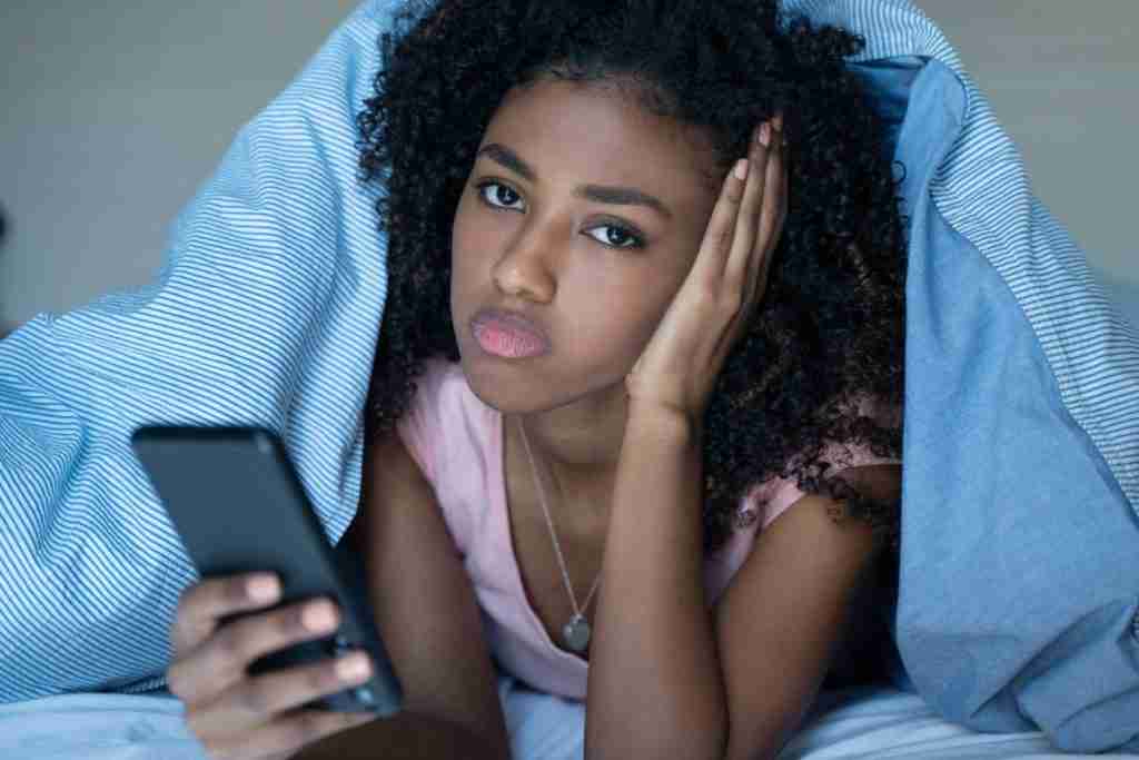 Black lady in bed under the cover holding her mobile phone - digital detox tips