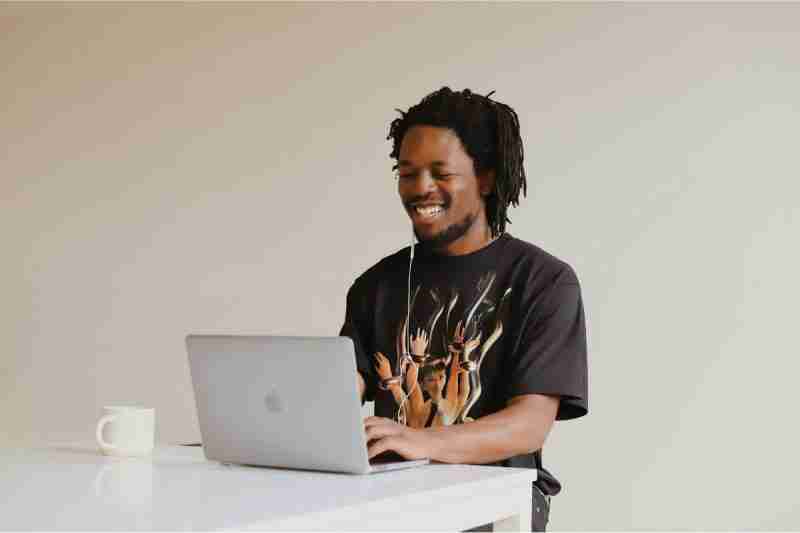 Black man in black tee shirt grinning with a broad smile showing white teeth as works on a laptop