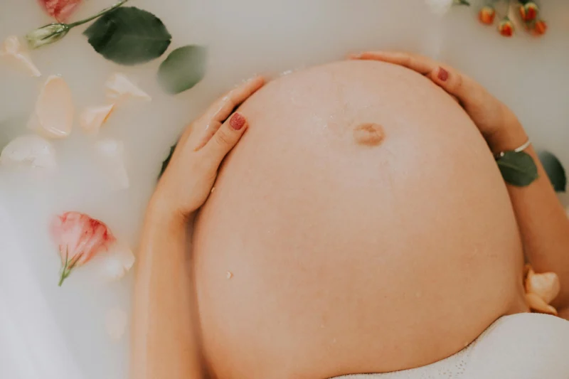 Close-up of a pregnant woman’s belly in a milk bath with rose petals and green leaves floating around, her hands gently resting on her stomach.