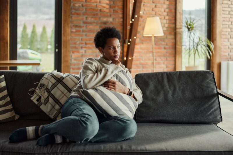 black lady in blue jeans seated on sofa and holding cushion to her abdomen contemplating NHS fibroids treatment