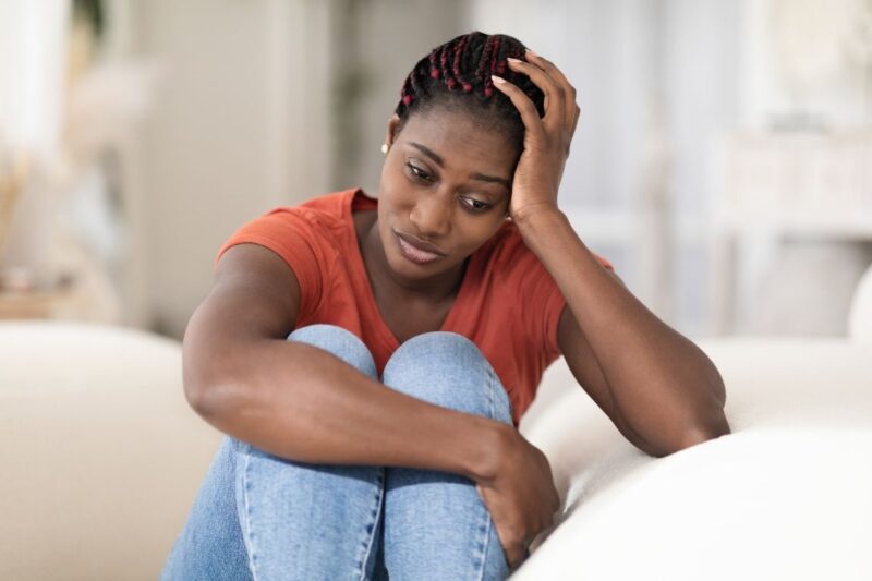 Upset pensive looking black woman  with hand to her head dressed in brown tshirt and blue jeans worried about vaginal lump 