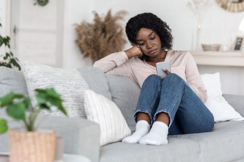Black woman pensively searching on her phone for answers to vaginal lumps that look like genital warts dressed in light pink blouse and blue jeans